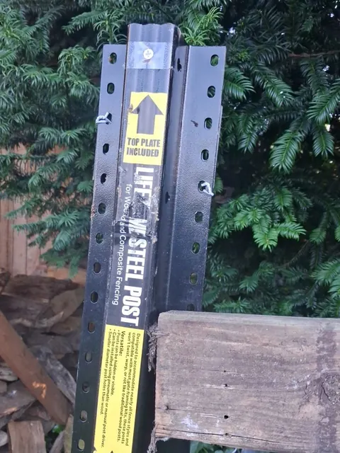 A dark metal fence-post component sits beside an existing wood rail, showing the connection area up close.