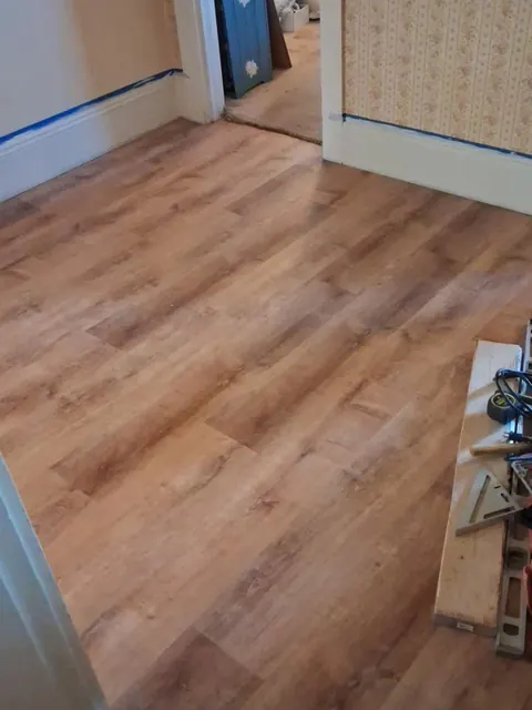 Wood-look flooring meets white baseboards and a doorway, with blue masking tape still visible on the trim.