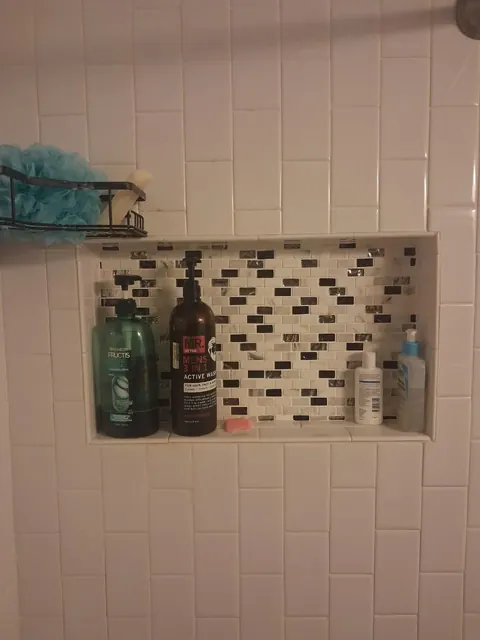 A mosaic-backed shelf is inset into a light tile shower wall, keeping bottles within reach.