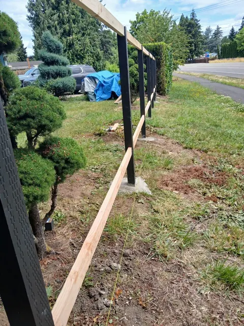 Dark posts and horizontal wood rails mark out a fence line beside the street before the infill is installed.