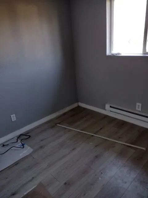 Gray walls, white baseboards and gray-brown plank flooring meet beneath a small window; supplies remain on the floor.
