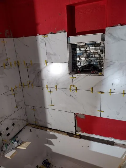Marble-look tiles and a mosaic niche are being fitted, with tile spacers and red-coated wall areas still visible.